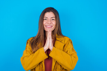 young brunette woman wearing yellow fringed jacket over blue background keeps palms together, has...