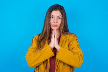 young brunette woman wearing yellow fringed jacket over blue background keeps palms pressed...