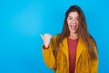 Fototapeta premium Stupefied young brunette woman wearing yellow fringed jacket over blue with surprised expression, opens eyes and mouth widely, points aside with thumb, shows something strange. Advertisement concept.