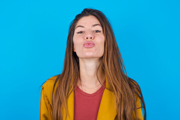 young brunette woman wearing yellow fringed jacket over blue background, keeps lips as going to kiss someone, has glad expression, grimace face. Standing indoors. Beauty concept.