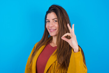 Glad attractive young brunette woman wearing yellow fringed jacket over blue background shows ok sign with hand as expresses approval, has cheerful expression, being optimistic.