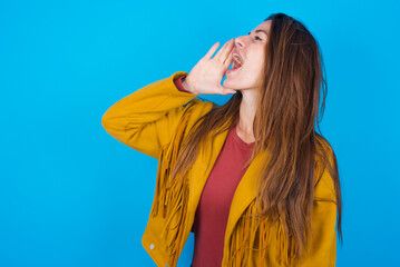 young brunette woman wearing yellow fringed jacket over blue background profile view, looking happy and excited, shouting and calling to copy space.