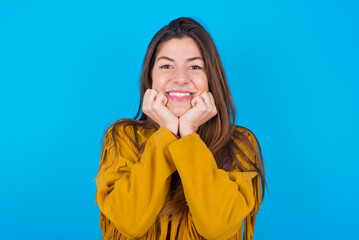 Dreamy young brunette woman wearing yellow fringed jacket over blue background keeps hands pressed...