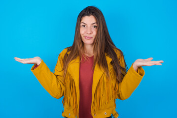 Puzzled and clueless young brunette woman wearing yellow fringed jacket over blue background with...