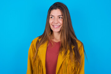 Oops! Portrait of young brunette woman wearing yellow fringed jacket over blue background clenches...