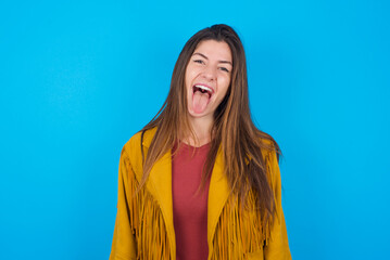 young brunette woman wearing yellow fringed jacket over blue background with happy and funny face...