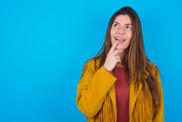 young caucasian woman wearing yellow fringed jacket over blue background with thoughtful...