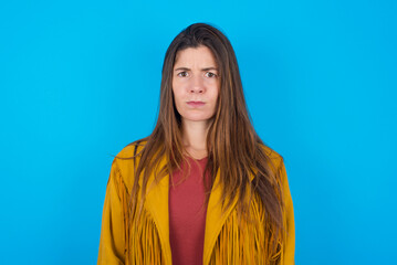 young caucasian woman wearing yellow fringed jacket over blue background frowning his eyebrows...