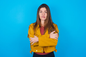 young caucasian woman wearing yellow fringed jacket over blue background bitting his mouth and...