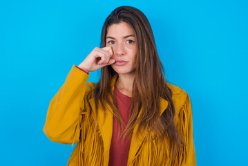 Unhappy young caucasian woman wearing yellow fringed jacket over blue background crying while...