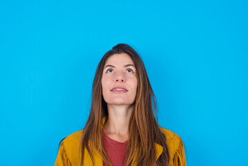 young caucasian woman wearing yellow fringed jacket over blue background looking up as he sees something strange.