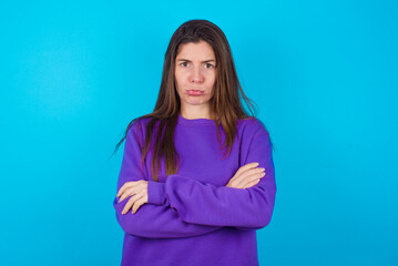 Obraz premium Gloomy dissatisfied young beautiful caucasian woman wearing purple sweater against blue background looks with miserable expression at camera from under forehead, makes unhappy grimace
