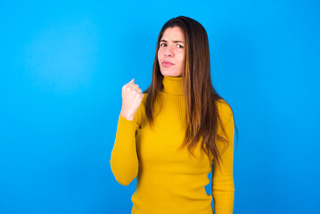 young woman wearing yellow turtleneck sweater against blue background shows fist has annoyed face...