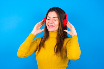 Pleased young woman wearing yellow turtleneck sweater against blue background enjoys listening pleasant melody keeps hands on stereo headphones closes eyes. Spending free time with music