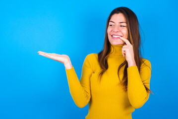 Positive young woman wearing yellow turtleneck sweater against blue background advert promo touches teeth with finger.