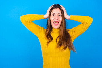 Cheerful overjoyed young woman wearing yellow turtleneck sweater against blue background reacts rising hands over head after receiving great news.