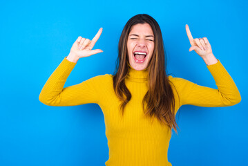 Photo of crazy young woman wearing yellow turtleneck sweater against blue background screaming and pointing with fingers at hair closed eyes