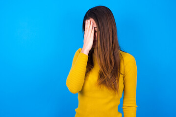 young woman wearing yellow turtleneck sweater against blue background with sad expression covering...