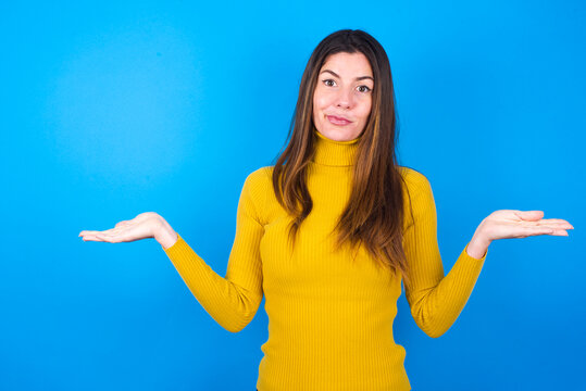 Careless attractive young woman wearing yellow turtleneck sweater against blue background shrugging shoulders, oops.