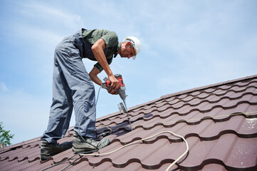 Worker prepearing to installation photovoltaic solar panel system on the roof of house. Man installer working with electric screwdriver, wearing uniform and helmet, on blue sky background.
