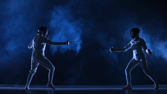 Two female athletes train on foil at fencing tournament. Young swordsmen in white uniforms and fencing masks fight duel on dark studio background with smoke and blue light. Fencing school. Slow motion