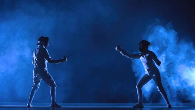 Two female athletes train on foil at fencing tournament. Young swordsmen in white uniforms and fencing masks fight duel on dark studio background with smoke and blue light. Fencing school. Slow motion