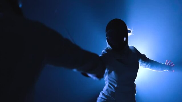 View from behind on two athletes fencers competing in tournament. Women in fencing masks attack each other, striking with epee. Dark studio with backlit blue light and smoke. Slow motion. Close up.