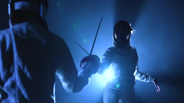 View from behind on two athletes fencers competing in tournament. Women in fencing masks attack each other, striking with epee. Dark studio with backlit blue light and smoke. Slow motion. Close up.