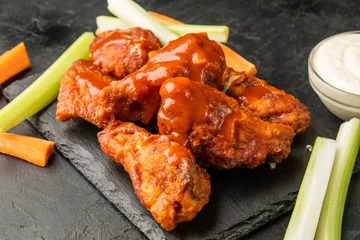 Foto auf Acrylglas BBQ wings. American recipe for buffalo wings in red sauce with vegetables cut into sticks, dark table. © Alexandr Milodan