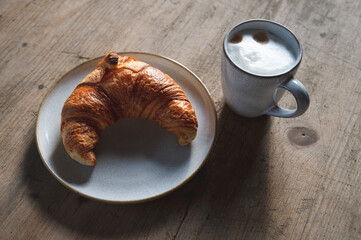 fresh golden brown croissant with a cappuccino on a stoneware plate and rustic wooden table