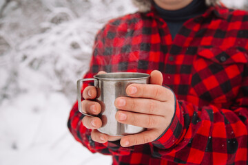 A hand holds a mug of hot tea. Drink tea in the winter outside. A metal cup with a drink is held by...