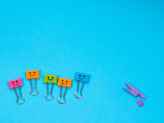 Funny multicolored smile metal binder clip or paper clip deride or laugh wooden violet clothespin with fracture leg on blue background. Copy space for text. Different, stand out of crowd concept