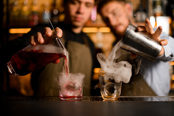 Obraz premium Two glasses stands on bar and two bartenders fills them with smoke and alcohol