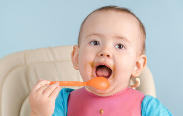 smiling newborn 12-17 months old in a pink bib puts a spoon with vegetable broccoli puree in his mouth