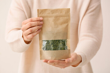 Woman holding paper bag with dried green tea leaves on light background, closeup