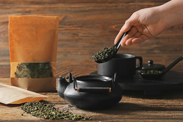 Woman pouring dried green tea leaves into teapot on wooden background