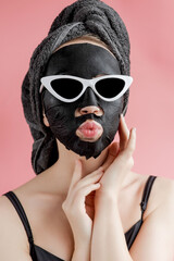 Young woman in glasses appling black cosmetic fabric facial mask on pink background. Face peeling mask with charcoal, spa beauty treatment, skincare, cosmetology. Close up