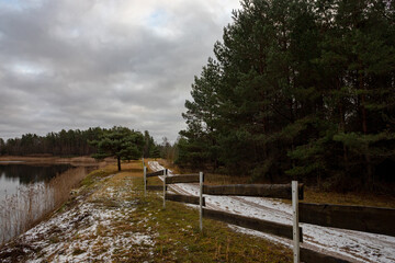 Fototapeta premium country road near pond, wooden fence, pine forest, first snow covered ground, cloudy sky, evening light