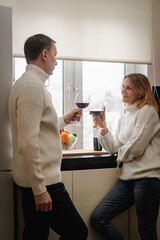 Romantic happy young couple in white clothes holding glasses of wine standing in modern cozy white kitchen, happy married husband and wife drinking red wine celebrate together at home