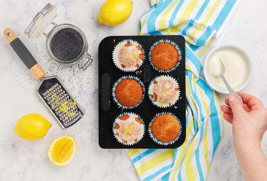 Women's hands decorate sugar icing on freshly baked cupcakes with poppy seeds and lemon zest on a marble background. The concept of home baking. Citrus dessert. Breakfast for gourmets