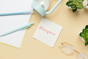 Sticky note with word MENOPAUSE, eyeglasses and pens on beige background
