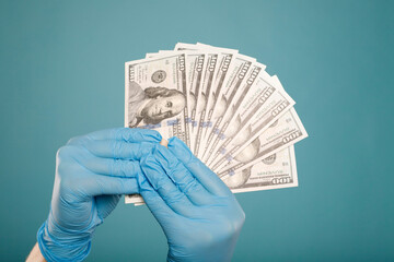 Close up of doctor's hands in blue protective gloves holding dollars over blue background....