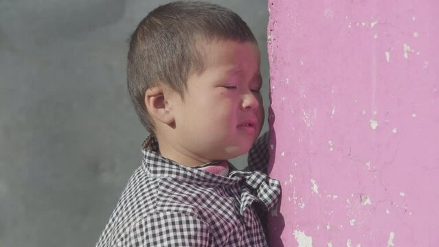 Asian little boy outside a pink brick house looking worried.