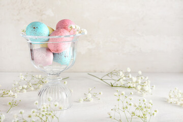 Glass bowl with painted Easter eggs and flowers on light background