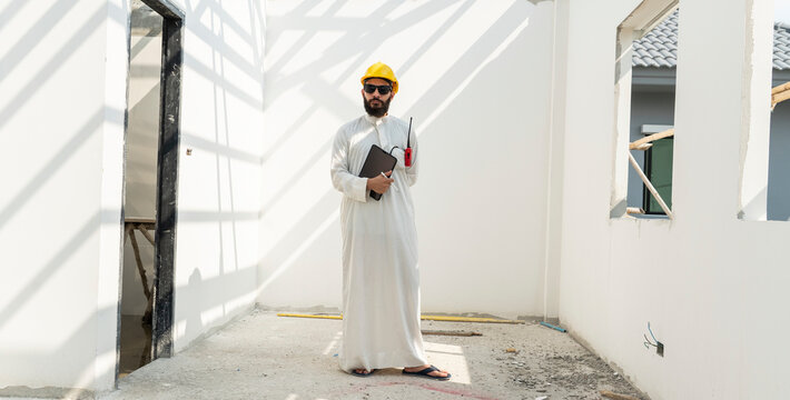 Middle Eastern Arab engineers dressed in traditional uniforms and safety helmets hold tablet and inspect construction projects in housing estates.Real estate development and property concept.