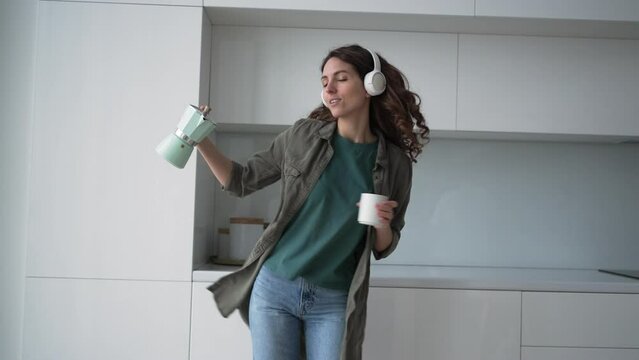 Curly-haired young woman with loose hairstyle dances to loud music in wireless headphones with coffee in cup and coffeepot slow motion