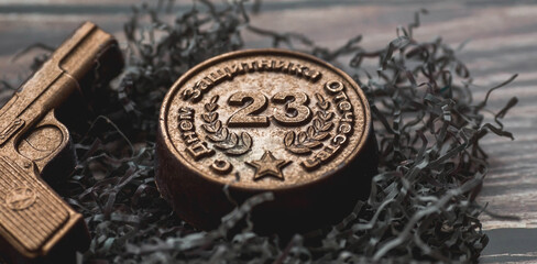 A holiday in Russia. Greeting card with Defender of the Fatherland Day, text in Russian. An iron medal with the date February 23 and a pistol on a wooden background. 
