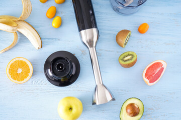 Black plastic electrical hand blender and accessories with sliced fruit on blue wooden background....