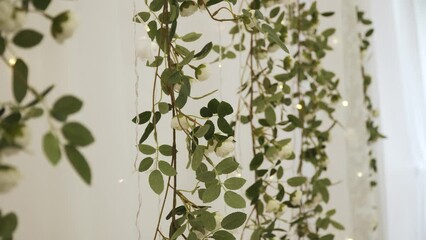 White wall decoration made of plastic garlands with green leaves and white rose flowers. Garlands of light bulbs hang down and blink yellow. - Powered by Adobe