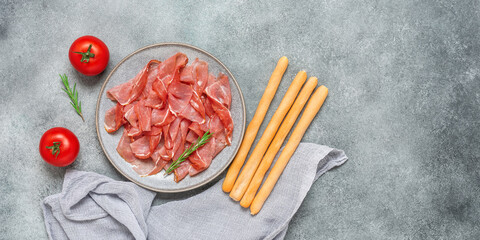 Slices of prosciutto di parma or jamon serrano in a plate and breadsticks on gray grunge background. Top view, flat lay, banner.